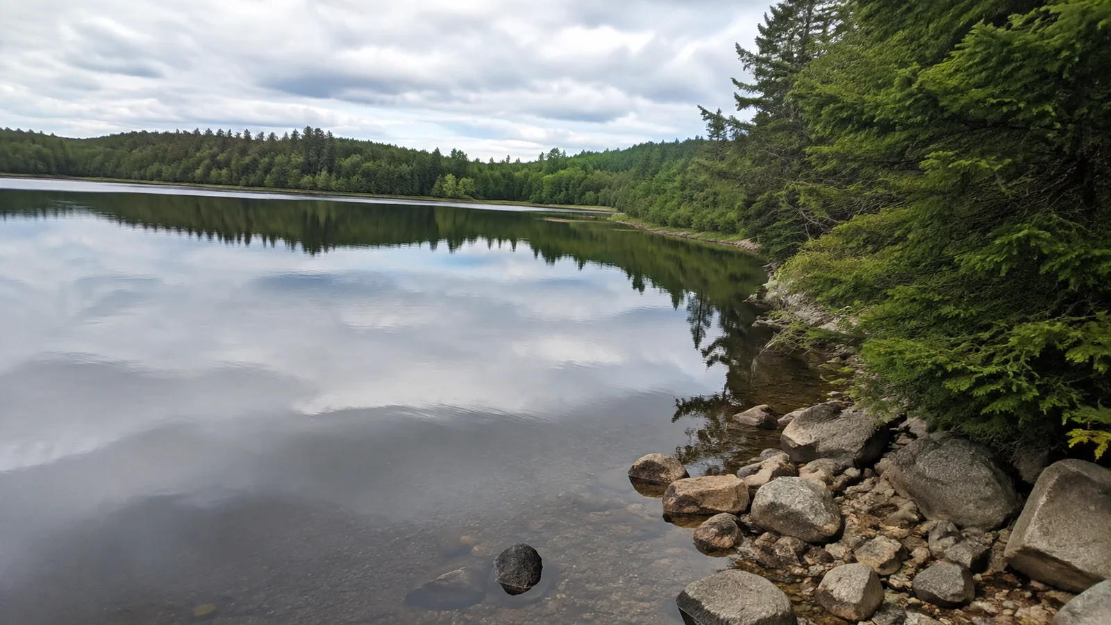 Still lake reflects clouds and trees beside a rocky shoreline.