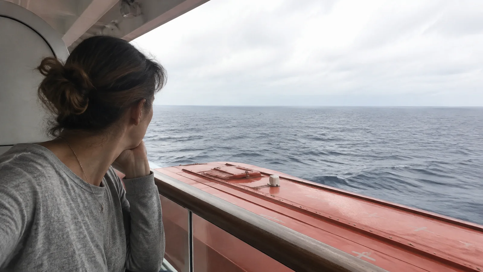 Passenger looking past an orange lifeboat from a sheltered deck.