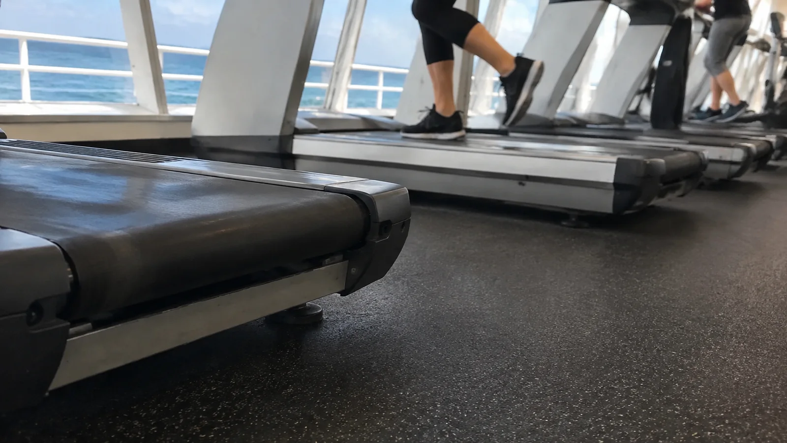 Passengers using treadmills beside ocean-facing gym windows.