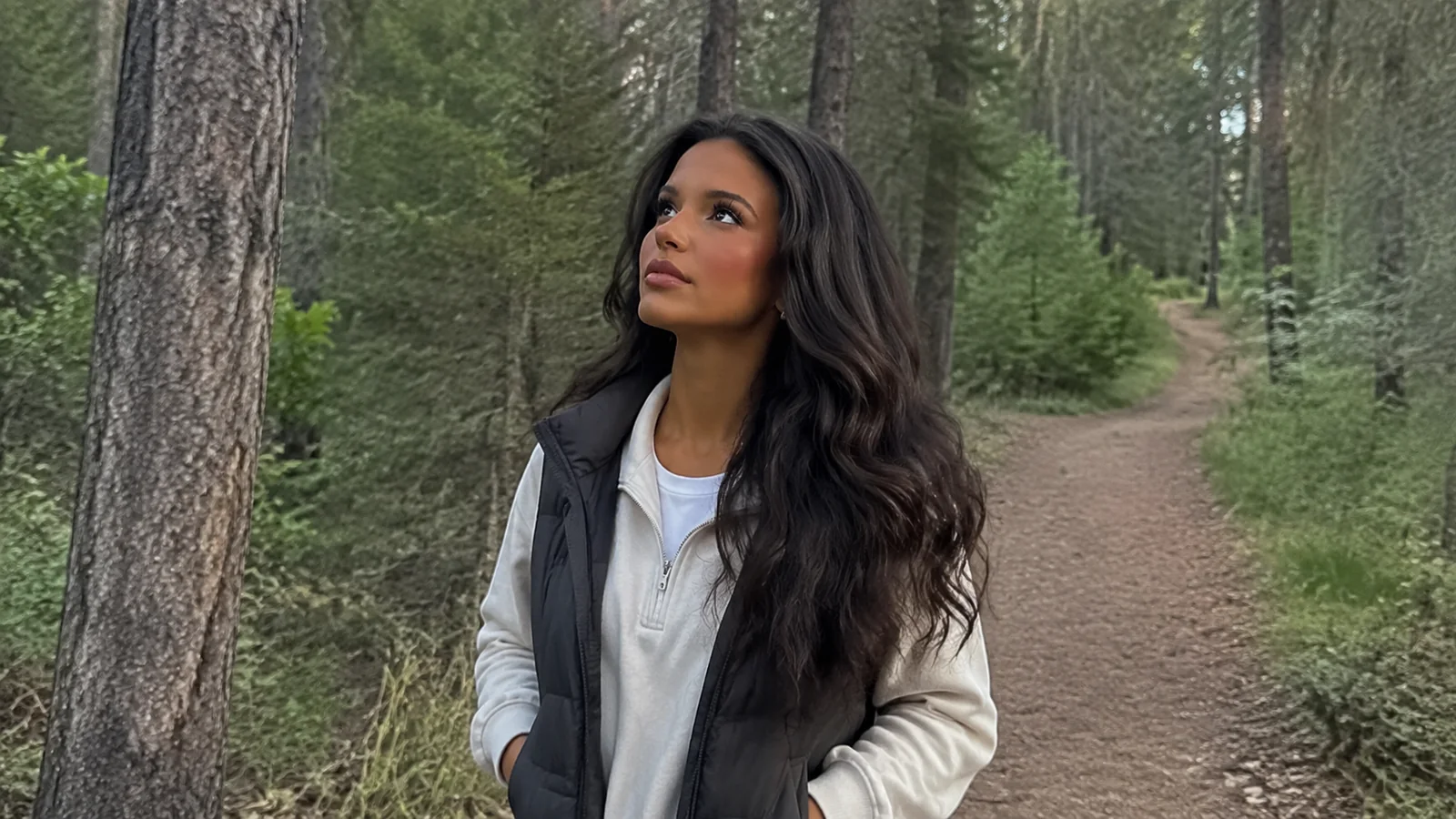 Woman looks up at tall trees along a forest path.