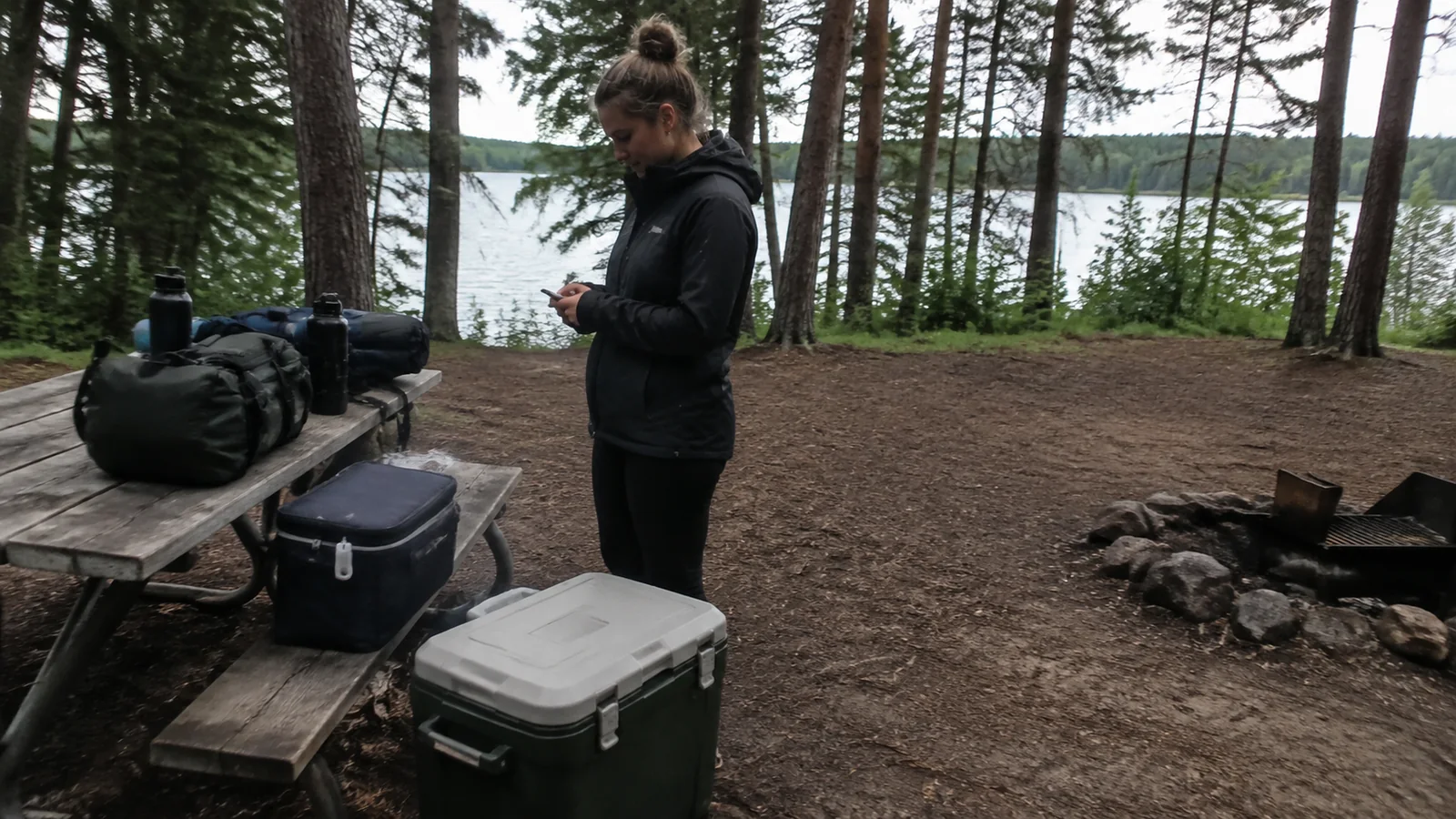 Camper stands beside coolers and bags at a wooded lakeside campsite.