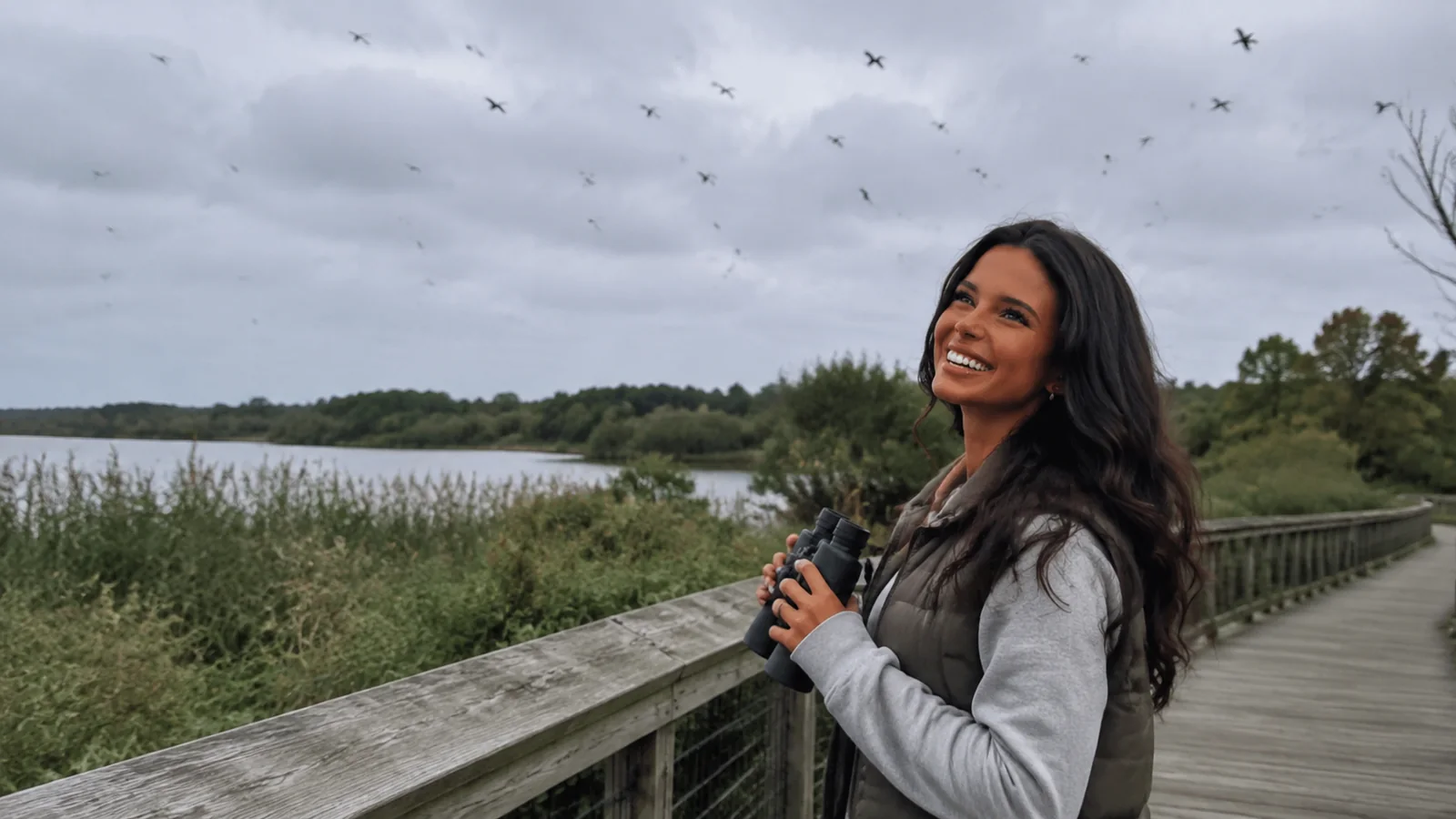 Woman holding binoculars on a wetland boardwalk with birds overhead.