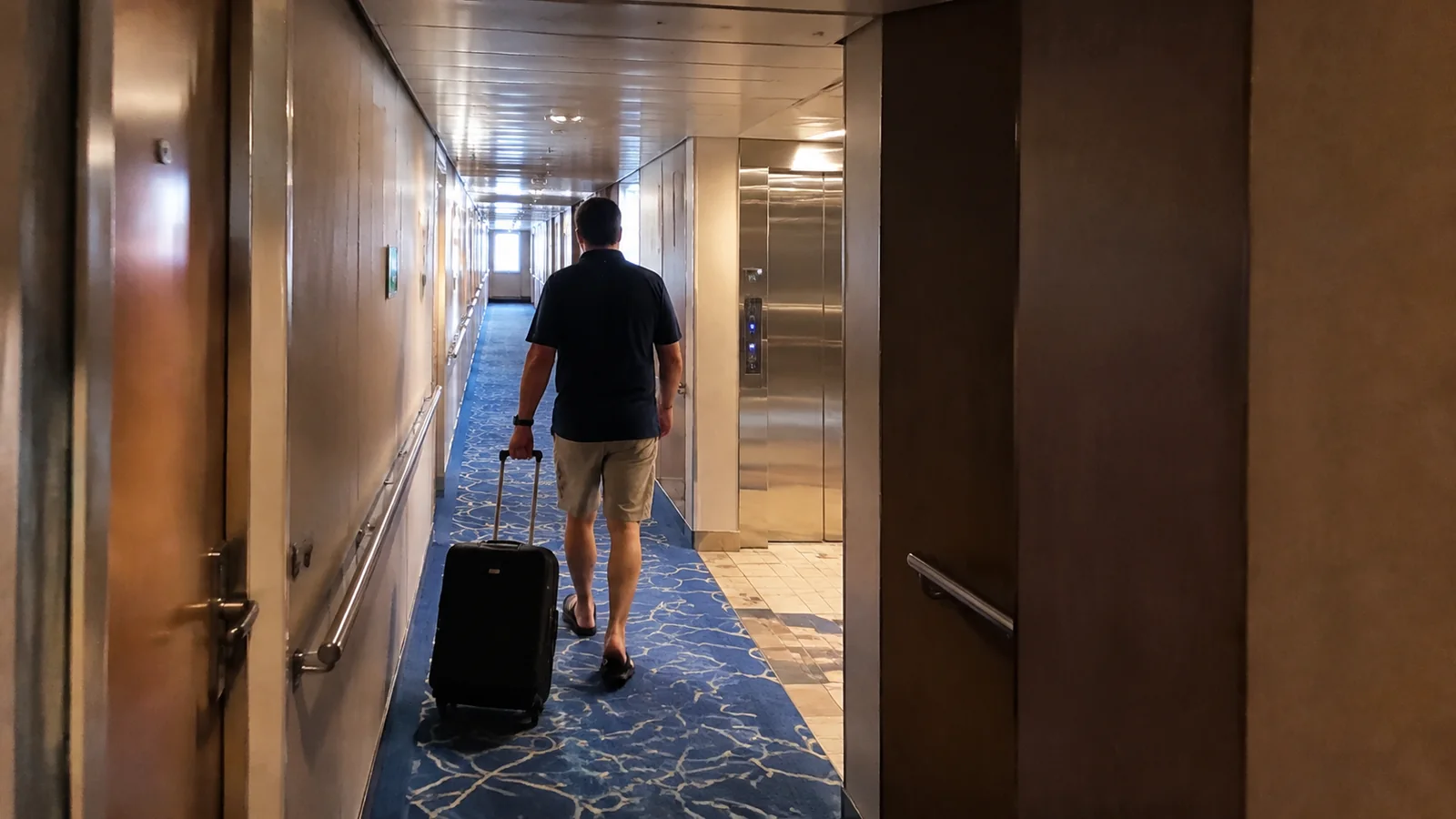 Man pulling a wheeled suitcase down a narrow ship corridor.