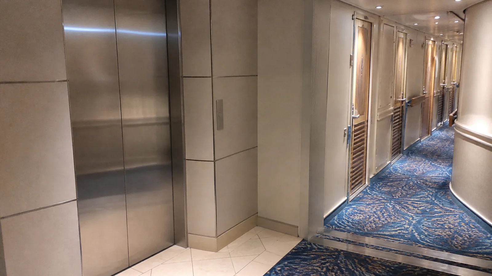 Ship elevator entrance beside a carpeted corridor lined with cabin doors.