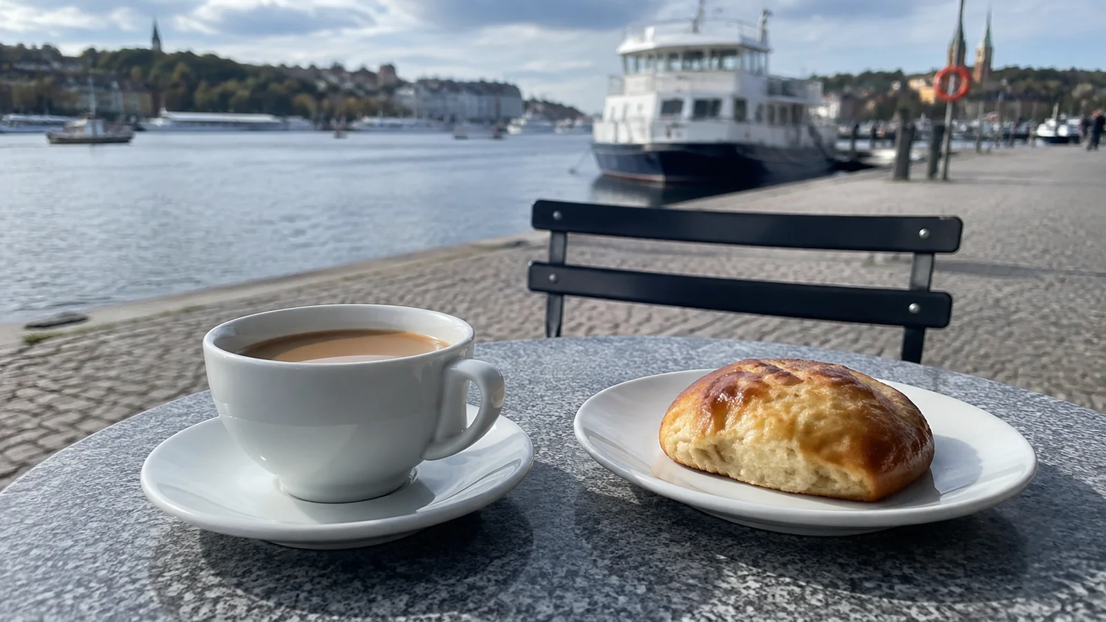 Coffee and a pastry on an outdoor table beside a harbor.