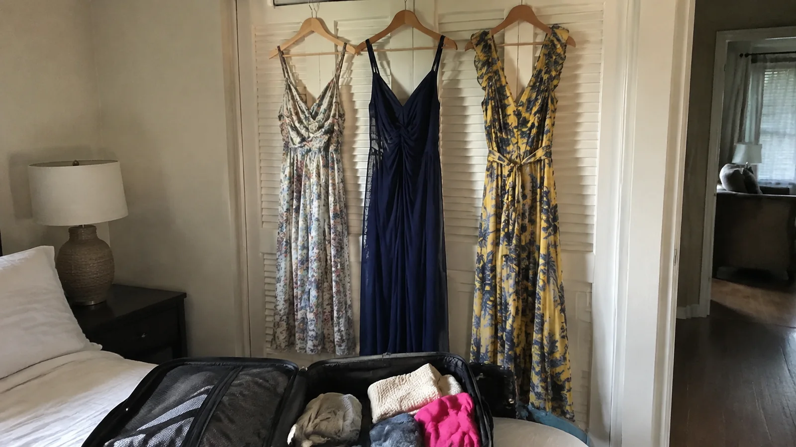 Three dresses hanging above an open suitcase beside a bed.