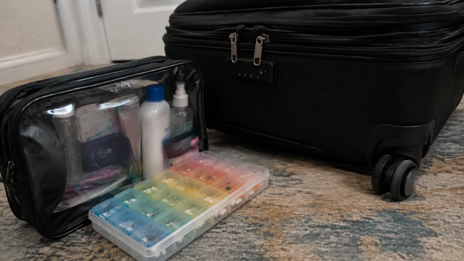 Toiletry pouch and pill organizer beside a carry-on suitcase.