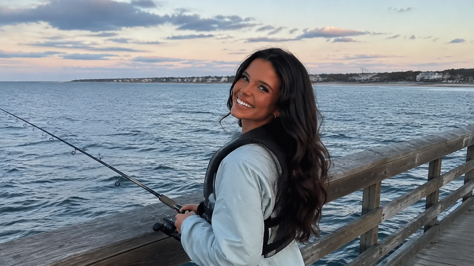 Woman holding a fishing rod beside a waterfront railing at sunset.