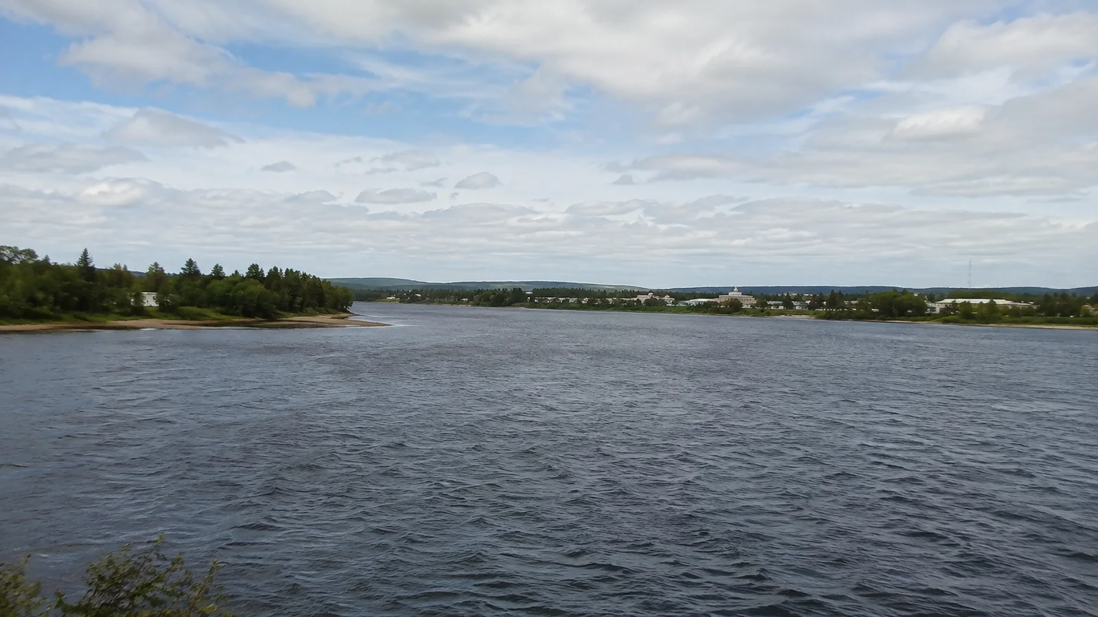 Wide lake with wooded shores and distant buildings under scattered clouds.