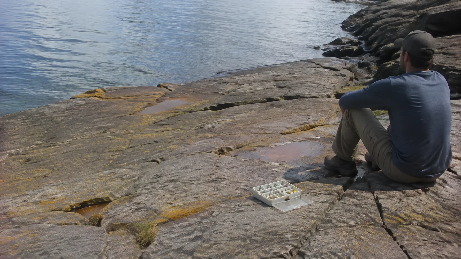 Angler sits on lakeside rocks beside an open tackle box.