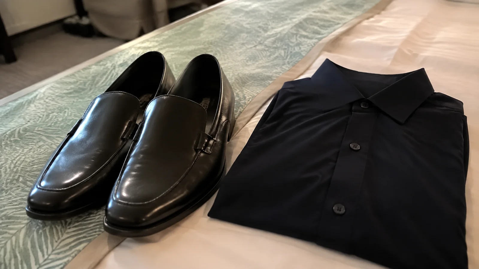 Black dress shoes and a folded dark shirt on a bed