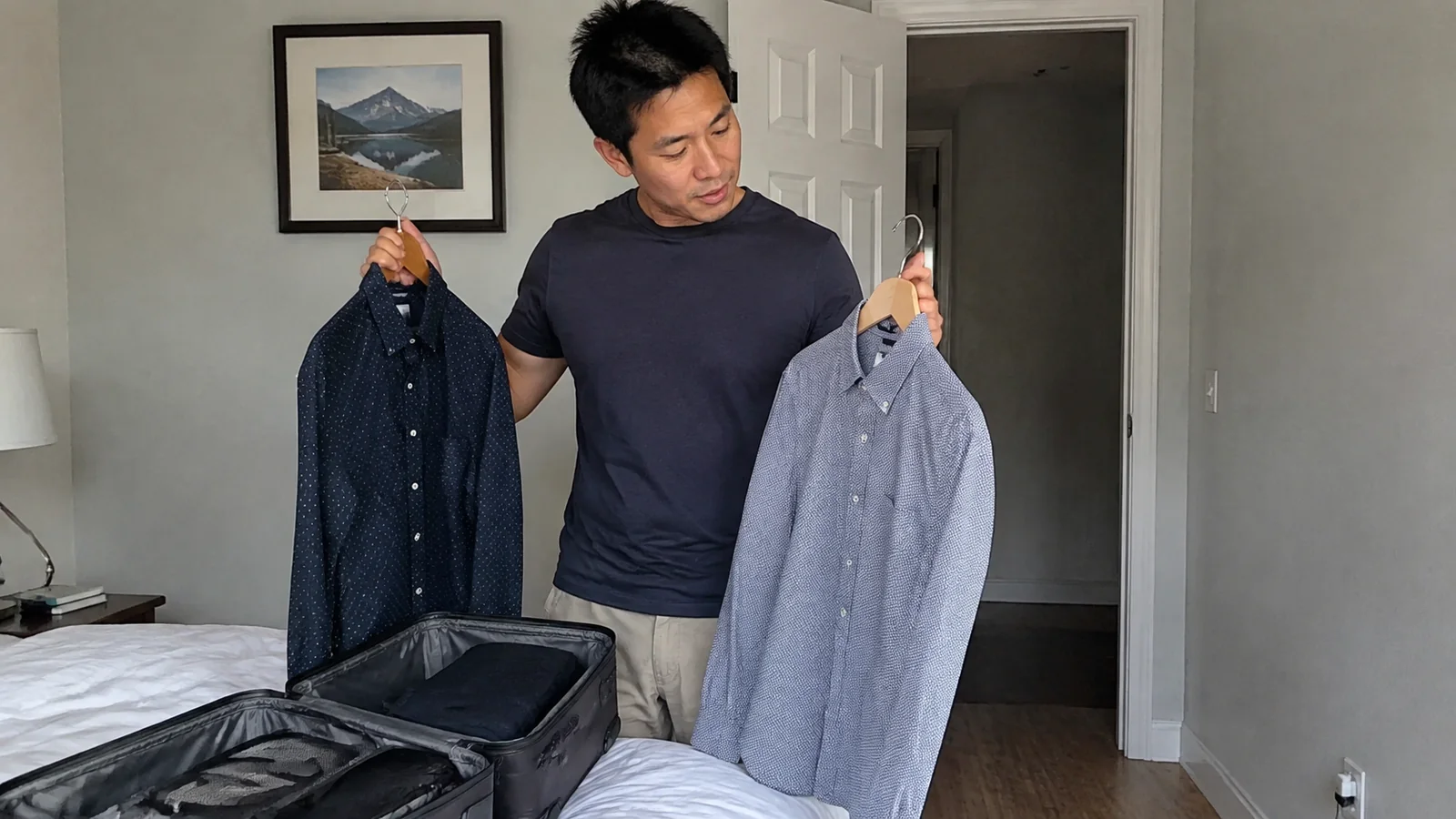 Man comparing two shirts on hangers beside an open suitcase