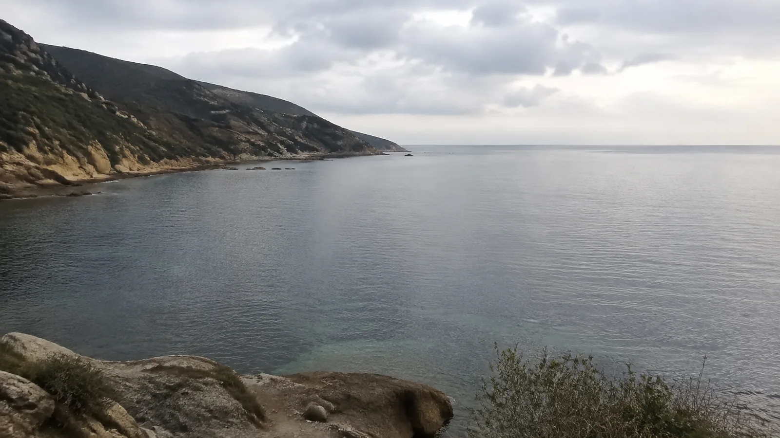 Calm coastal bay beneath rocky hills and an overcast sky