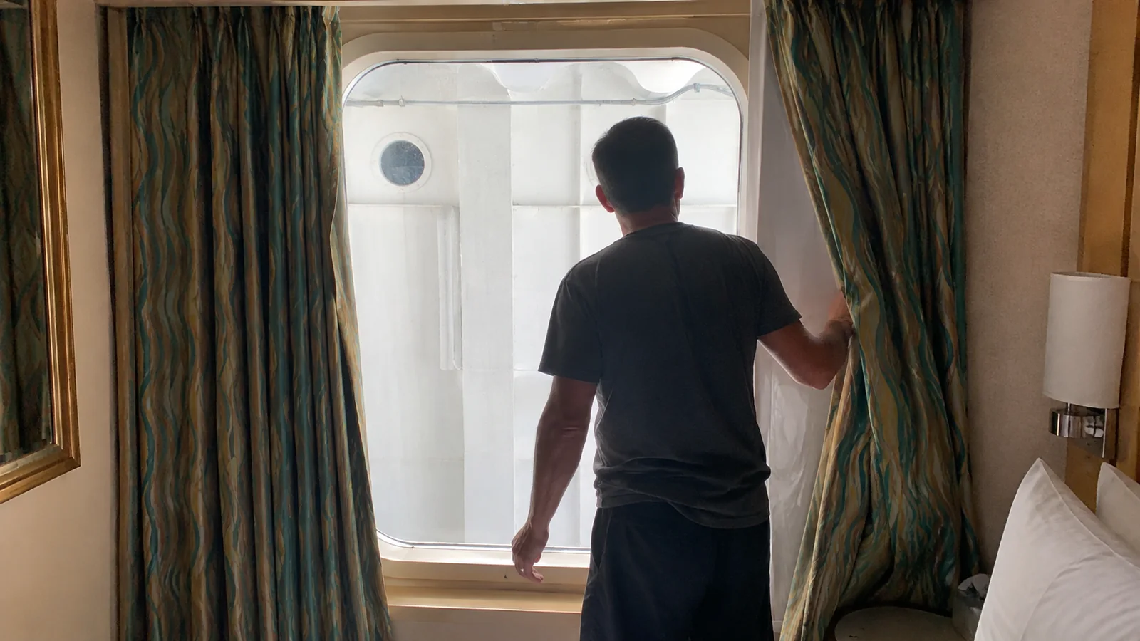Man looking through a cruise cabin window blocked by an exterior ship structure