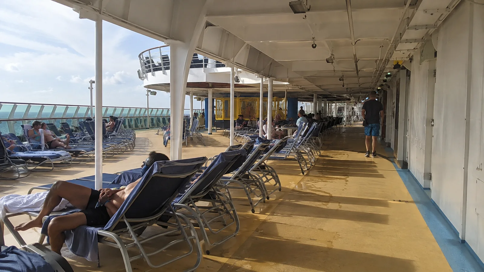 Passengers relaxing on lounge chairs along a sunlit cruise ship promenade