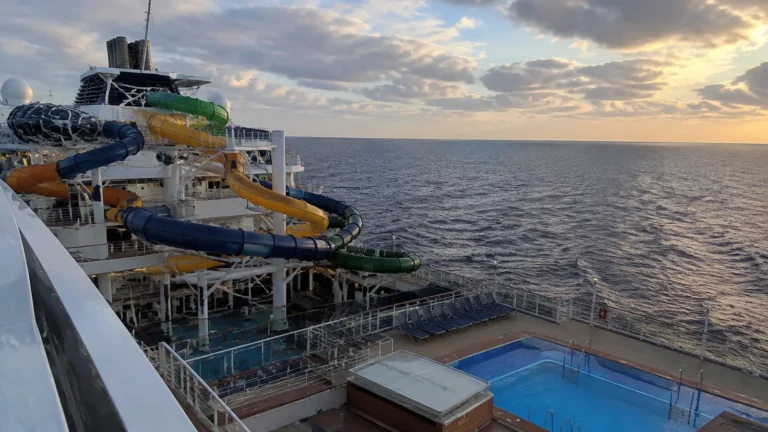 Colorful waterslides overlook a ship's pool and the ocean at sunset.