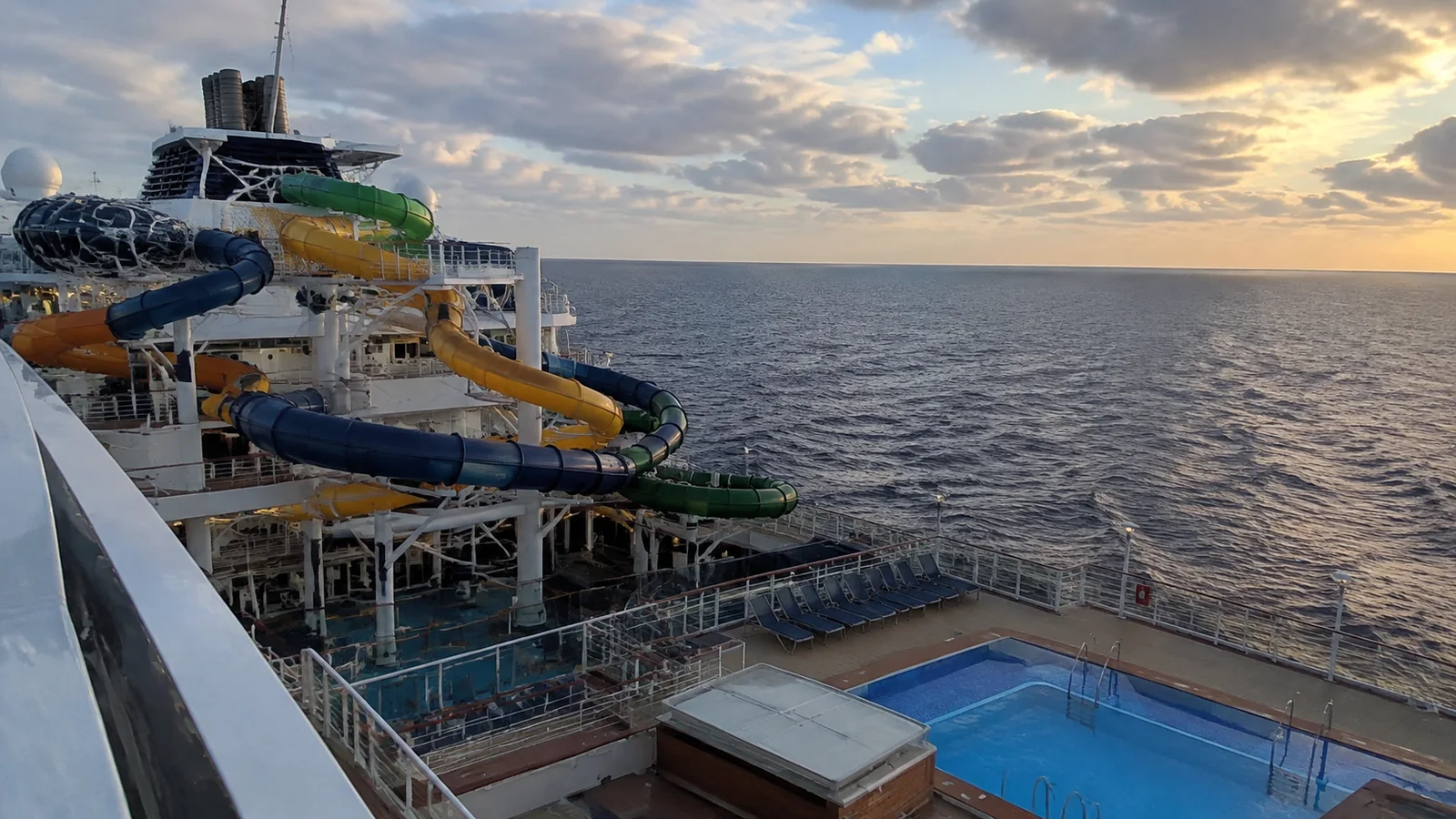 Colorful waterslides overlook a ship's pool and the ocean at sunset.