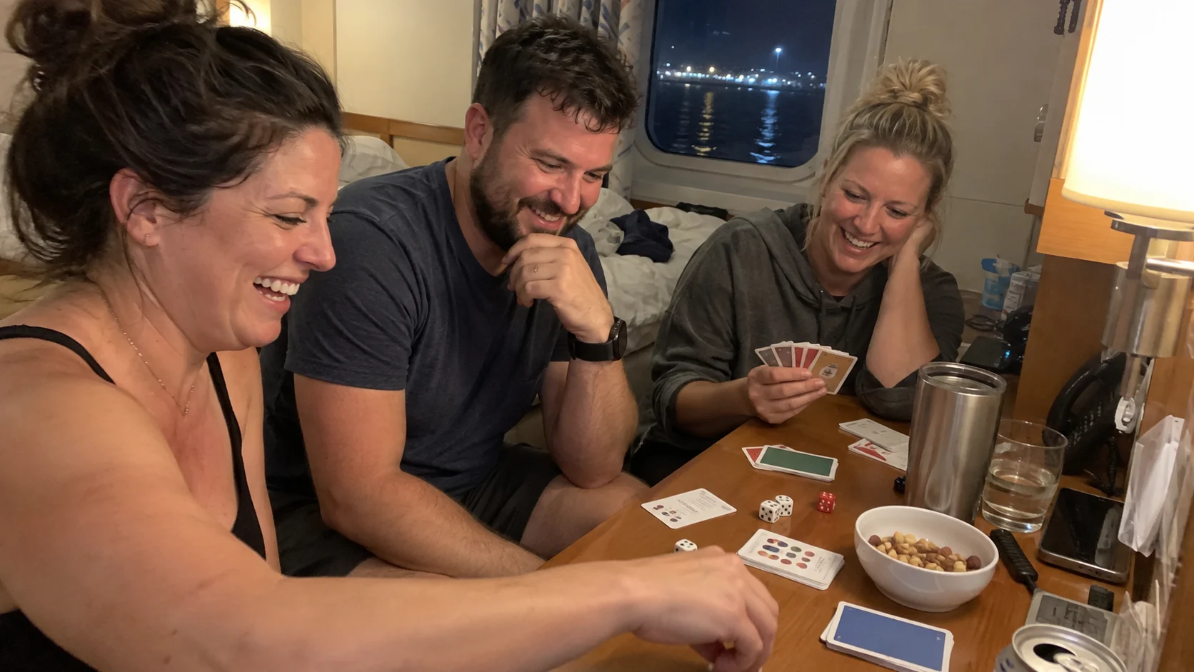 Three friends laughing over a pocket-size card and dice game inside a cruise cabin.