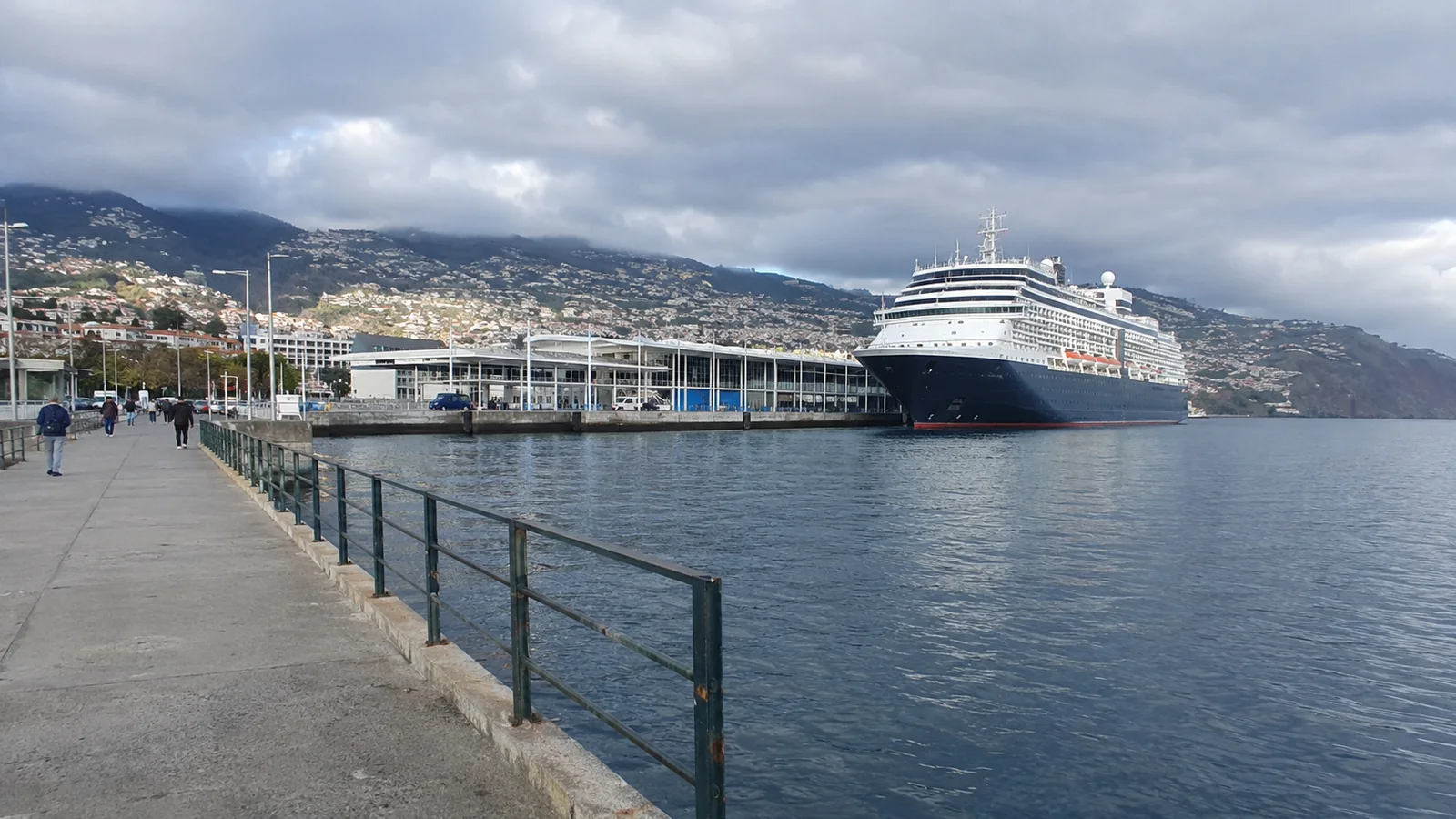Cruise ship docked beside a waterfront promenade with a hillside city behind