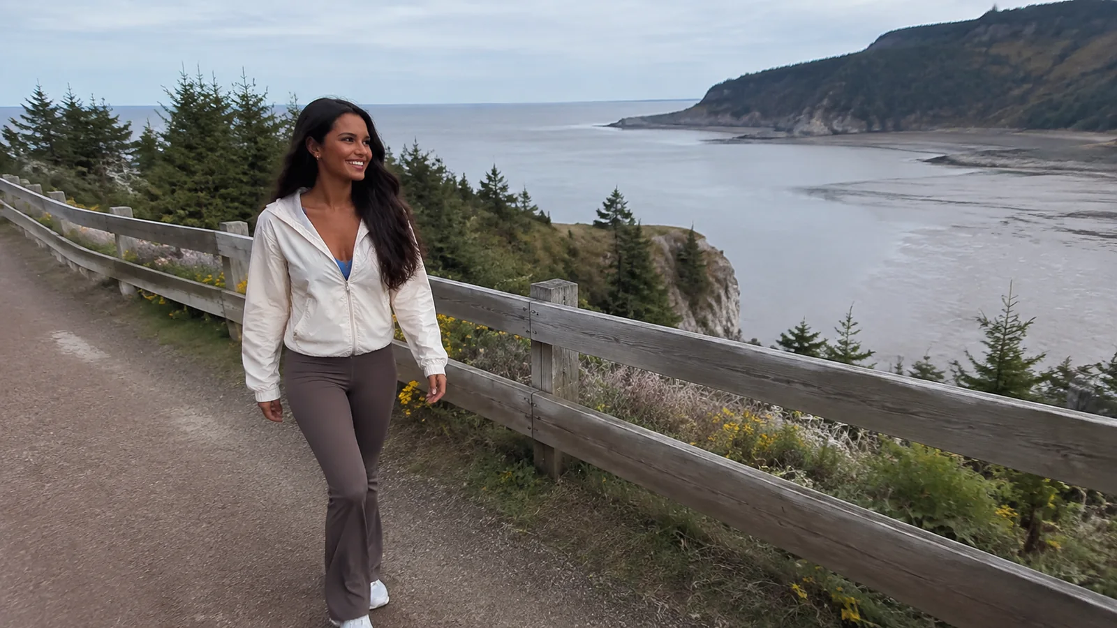 Woman walking a fenced coastal path overlooking a broad tidal bay.