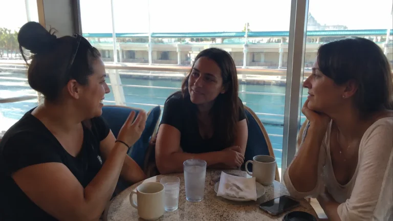 Three women chatting over coffee at a waterfront table.
