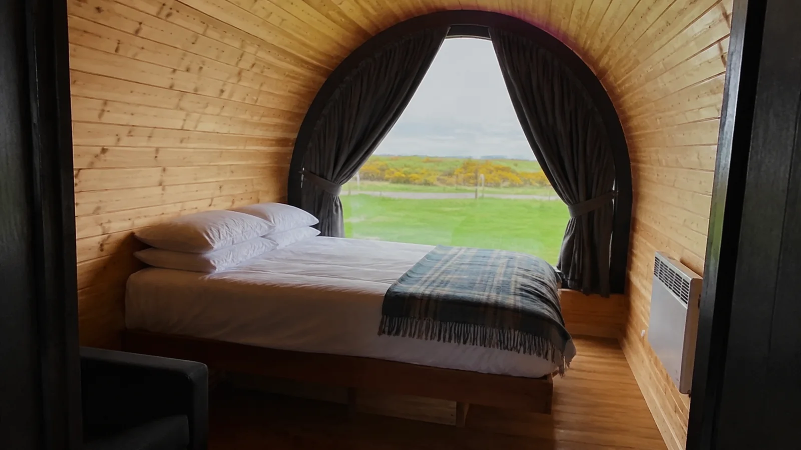 Bed inside a curved wood-paneled camping cabin with open curtains.