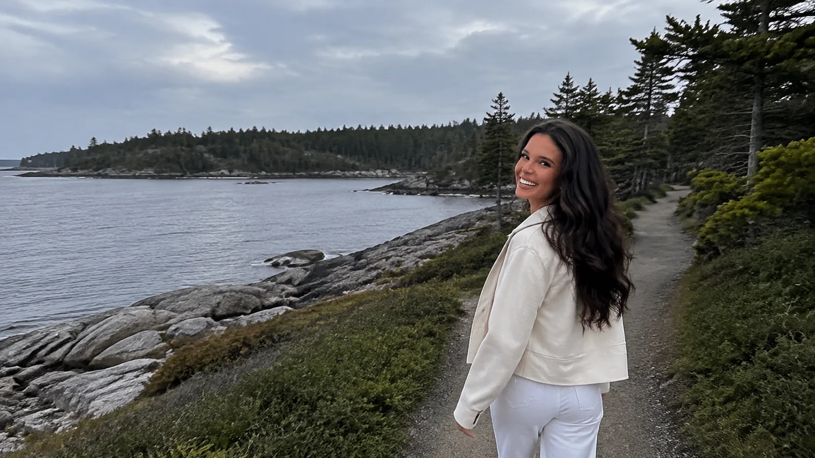 Woman walking a narrow coastal path above rocks and calm water.