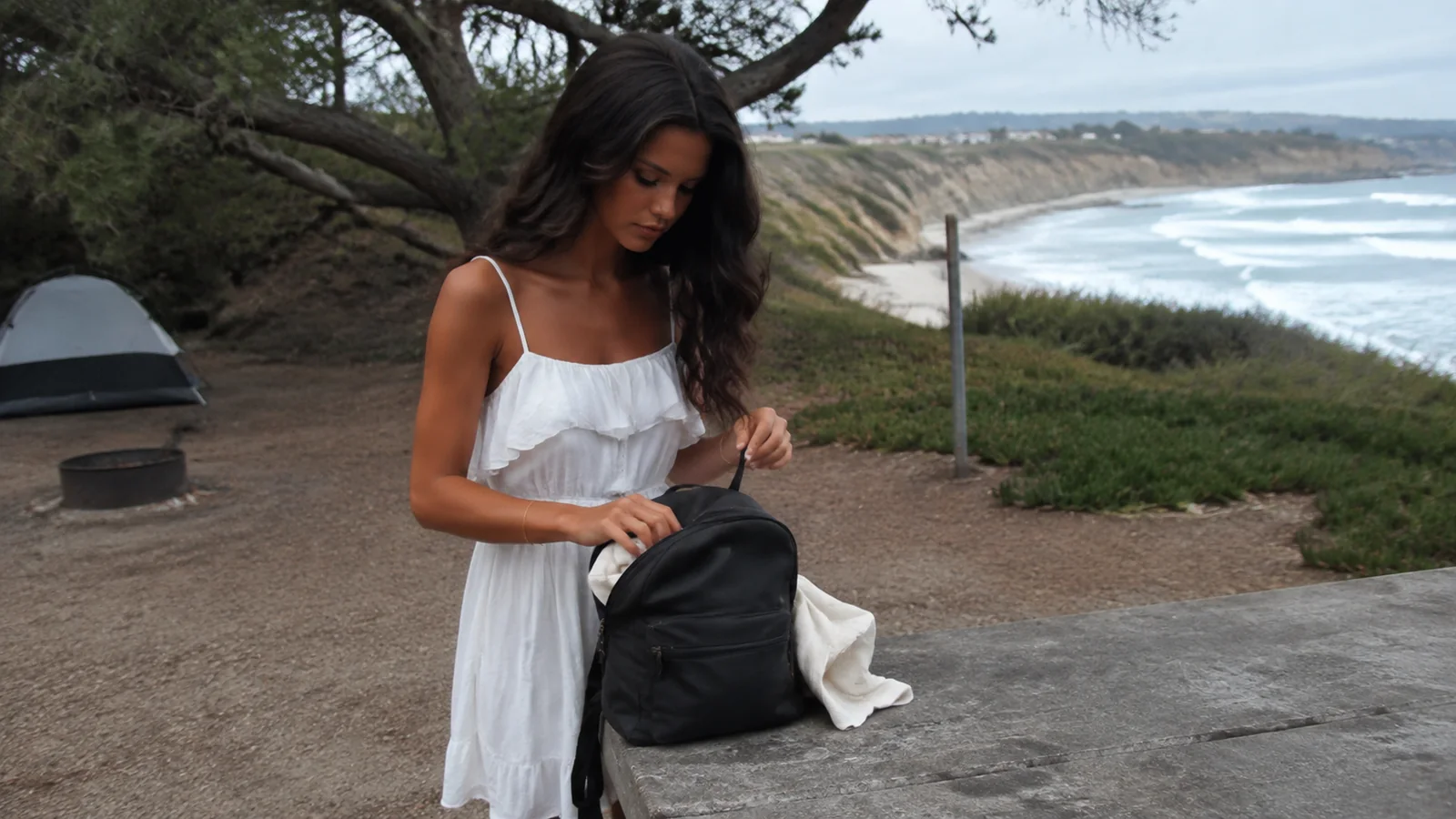 Woman packing a backpack at a seaside campsite with a tent behind.