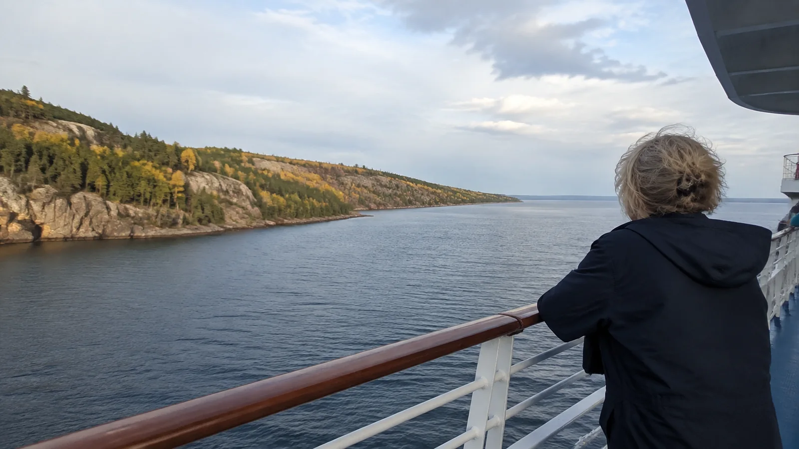 Passenger watching an autumn-colored rocky shoreline from an open deck.