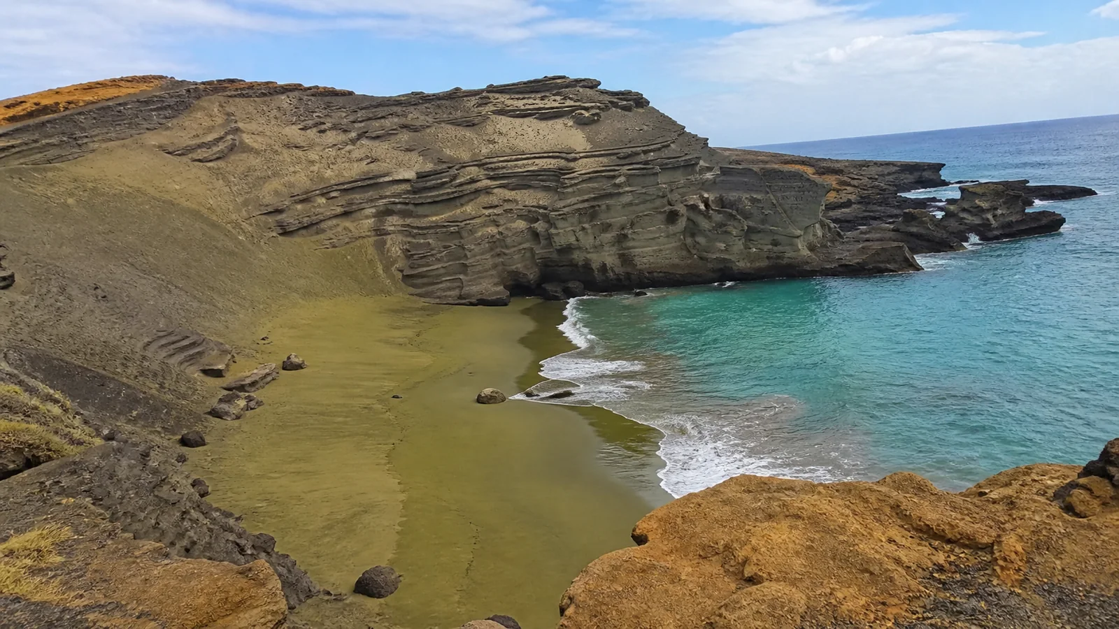 Turquoise water laps a green sand beach beneath layered coastal cliffs.