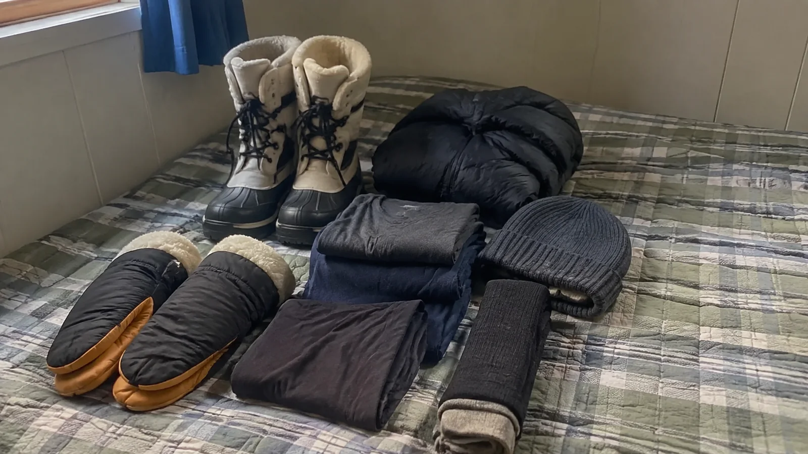 Winter boots, gloves, hat, and folded clothing arranged on a bed.