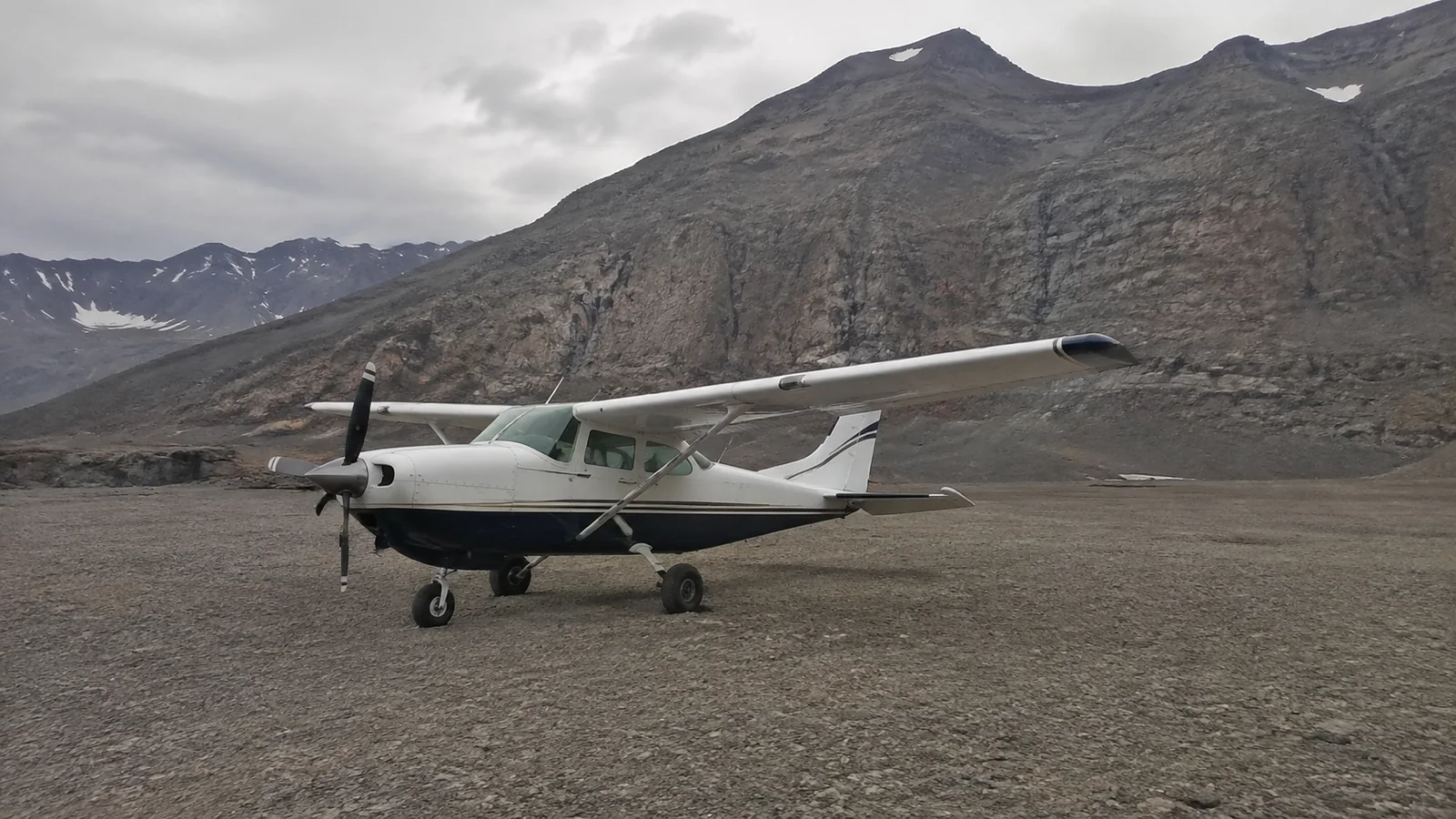 Small propeller airplane parked on a gravel airstrip below rocky mountains.
