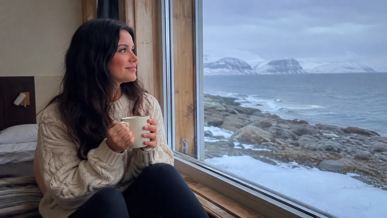 Woman holding a mug beside a window overlooking a snowy rocky coast.