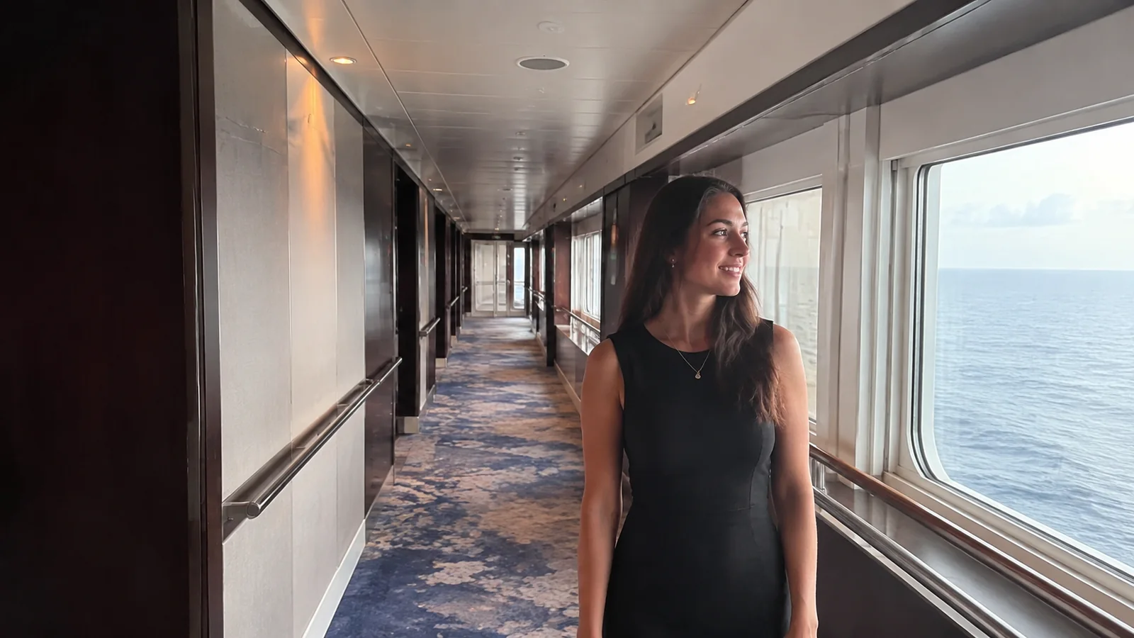 Woman walks along a ship corridor beside large ocean windows.