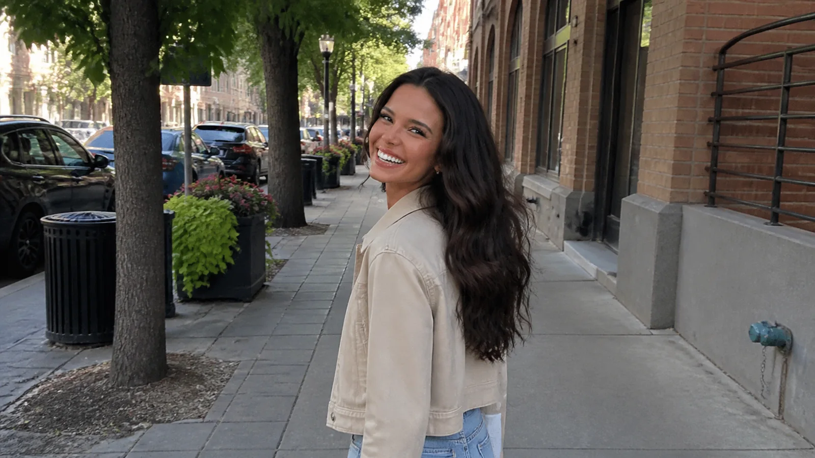 Woman walking a downtown sidewalk lined with trees and brick buildings.