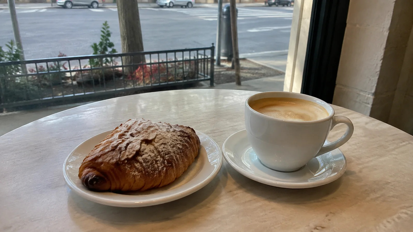 Coffee and a flaky pastry on a cafe table beside a window.