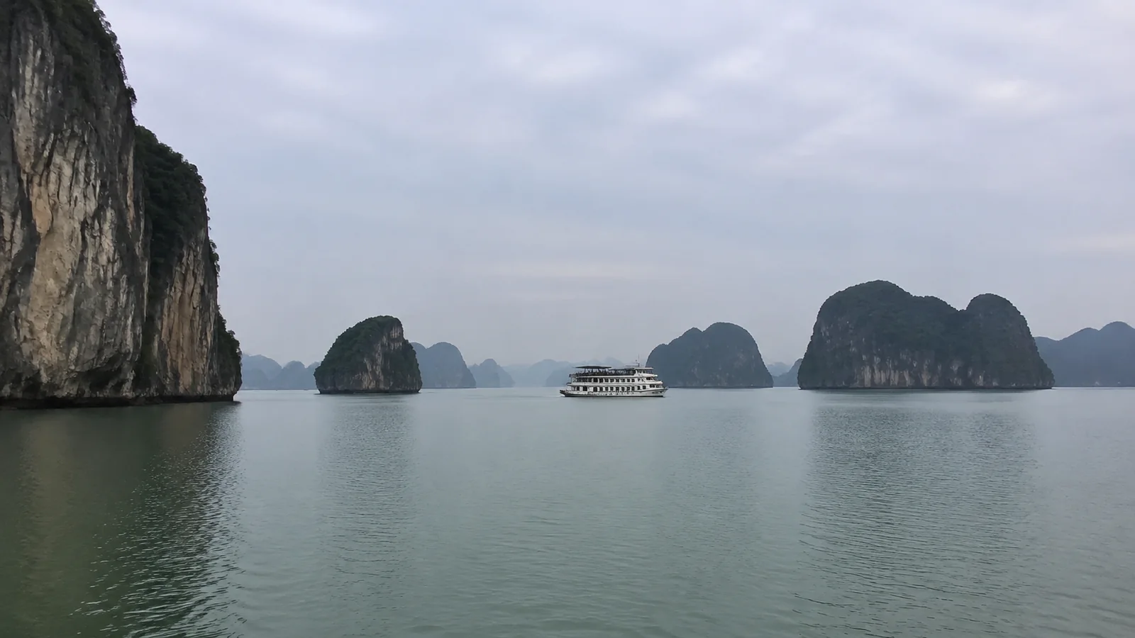 Cruise boat sailing among steep rocky islands beneath an overcast sky.
