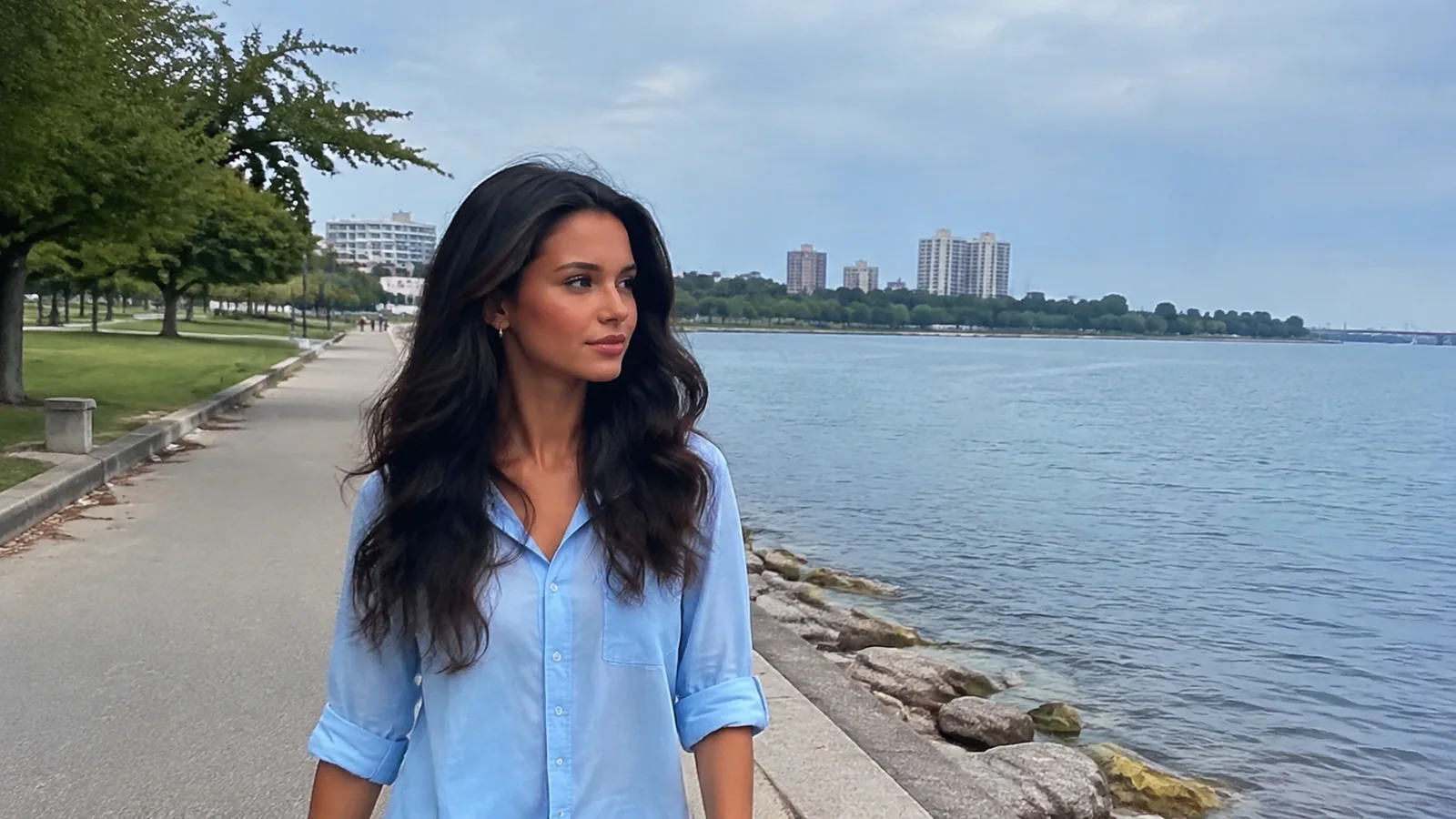 Woman stands on a lakeside path with distant buildings across the water.