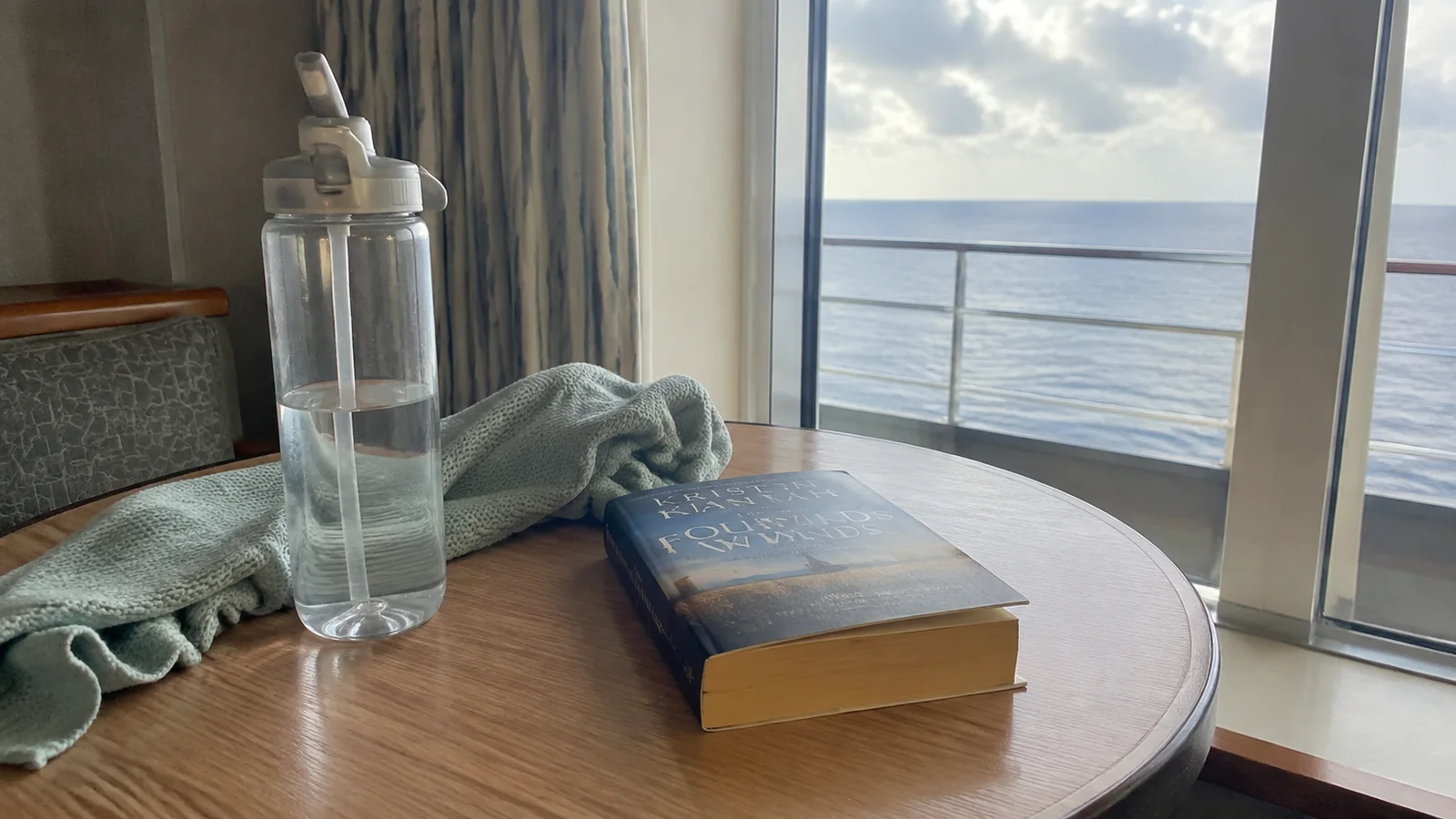Water bottle, book, and folded clothing on a cruise cabin table beside a balcony
