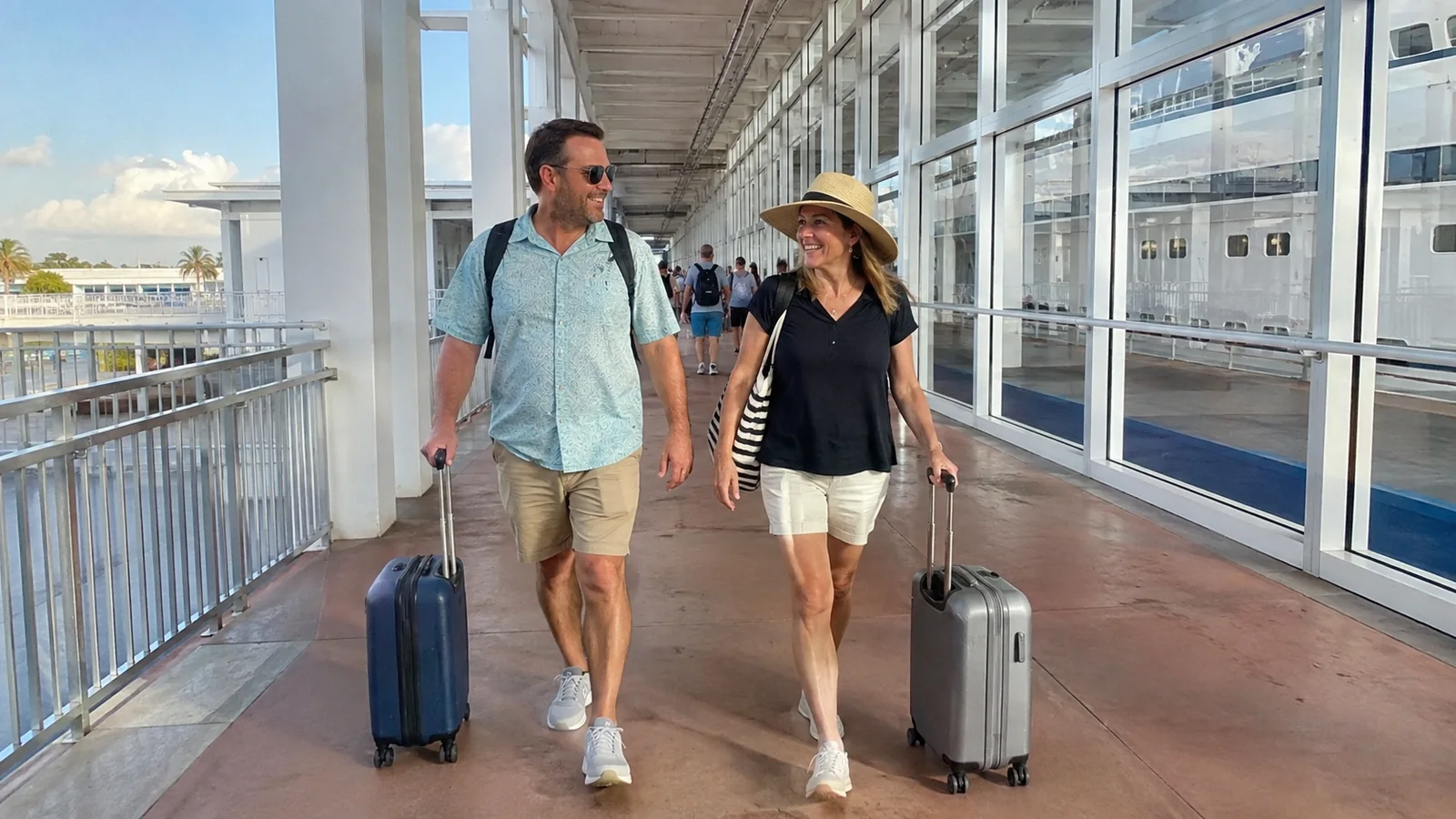 Couple pulling wheeled suitcases along a covered cruise terminal walkway.