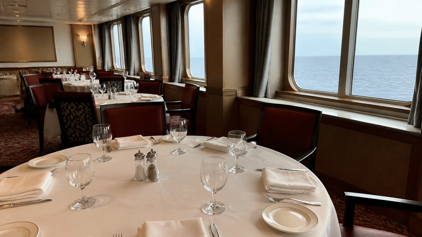 Round dining table set with white linens and glassware beside ocean windows.