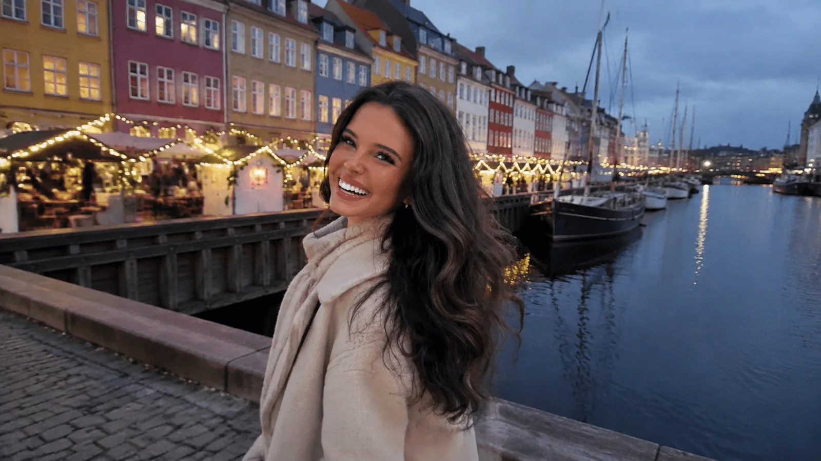 Woman walking beside a canal lined with festive market lights.