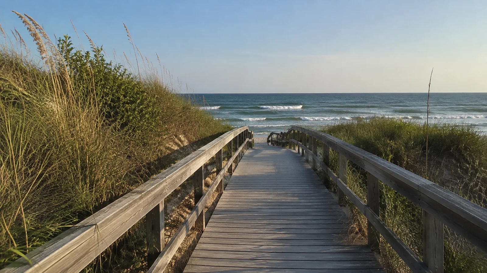 Wooden boardwalk descends between grassy dunes toward breaking waves.