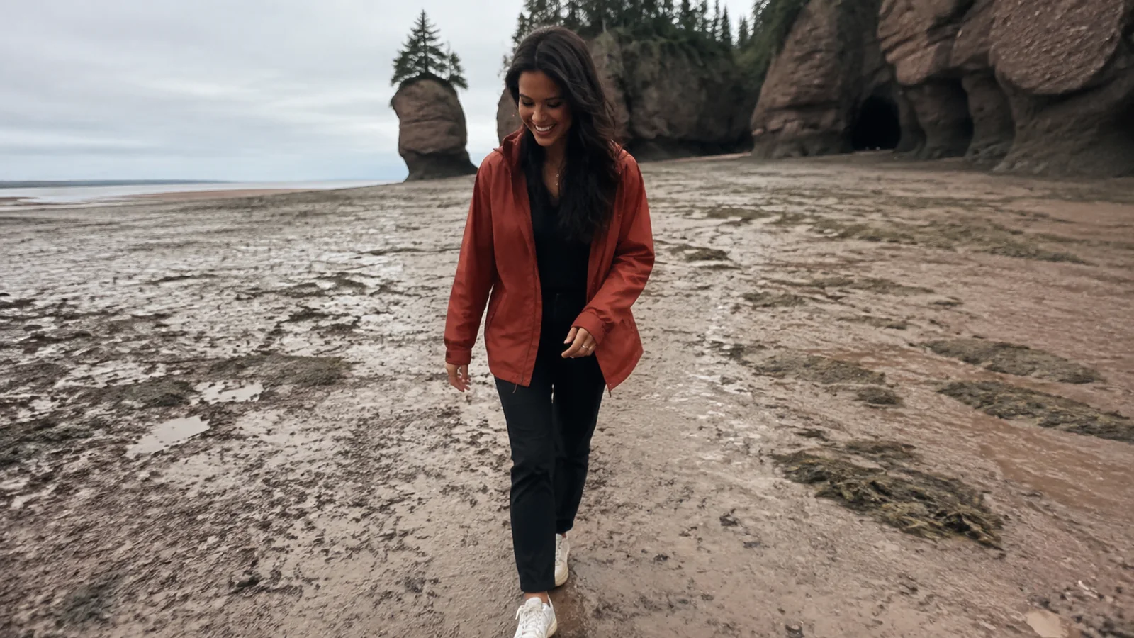 Woman walks across wet sand among towering coastal rock formations.