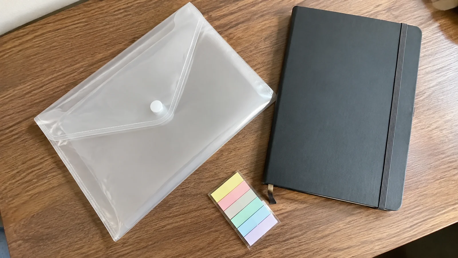 Clear document pouch, dark notebook and colored tabs lie on a wooden table.