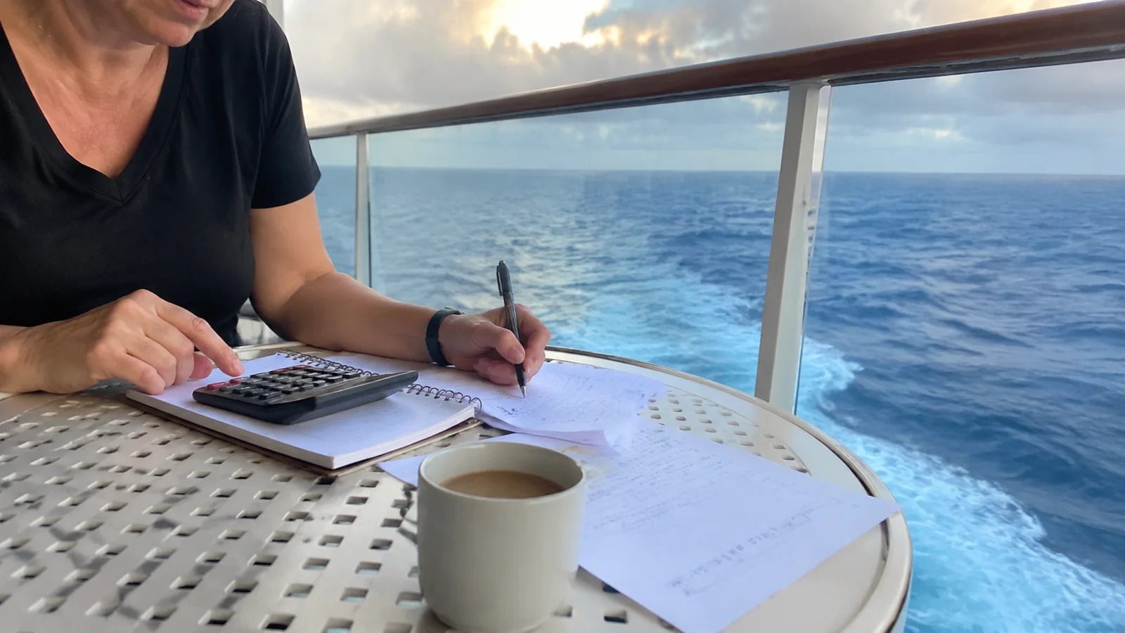 Woman using a calculator and writing in a notebook at a cruise ship balcony table