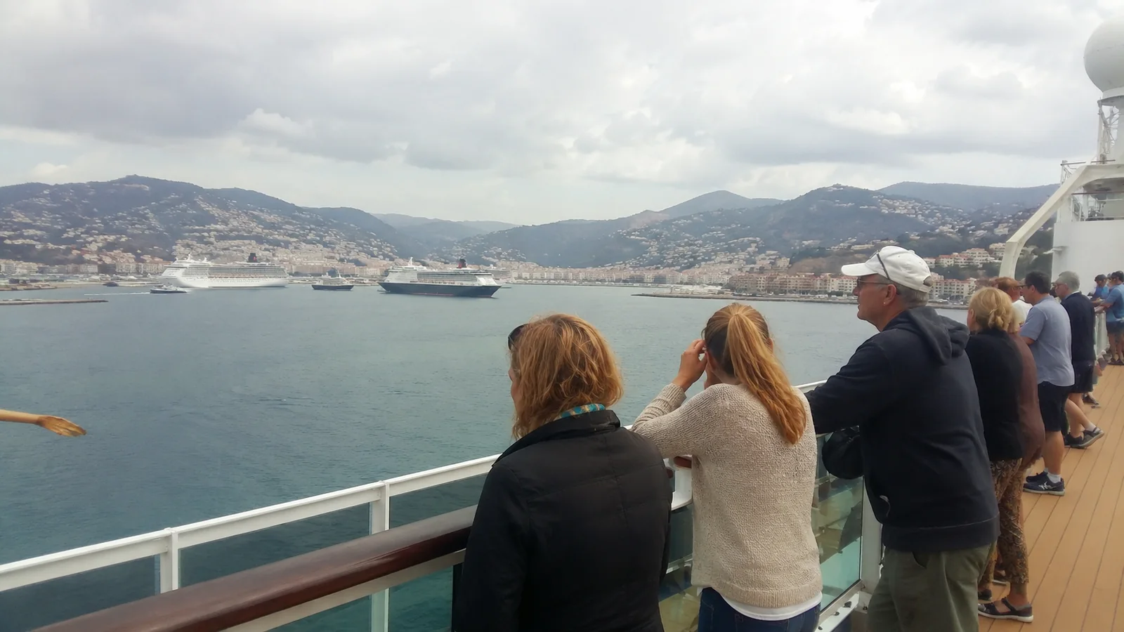Passengers standing at a cruise ship railing overlooking a harbor and mountain town