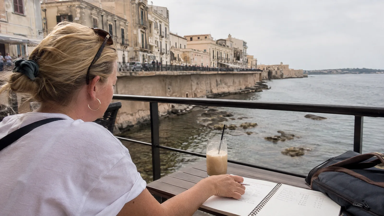 Woman writing in a notebook at a waterside table overlooking a historic coastal town