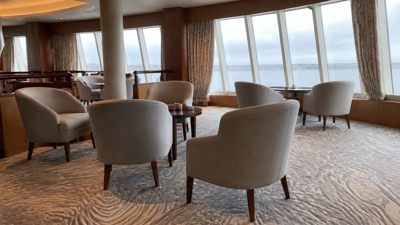 Empty lounge chairs grouped around small tables beside ocean-facing windows.