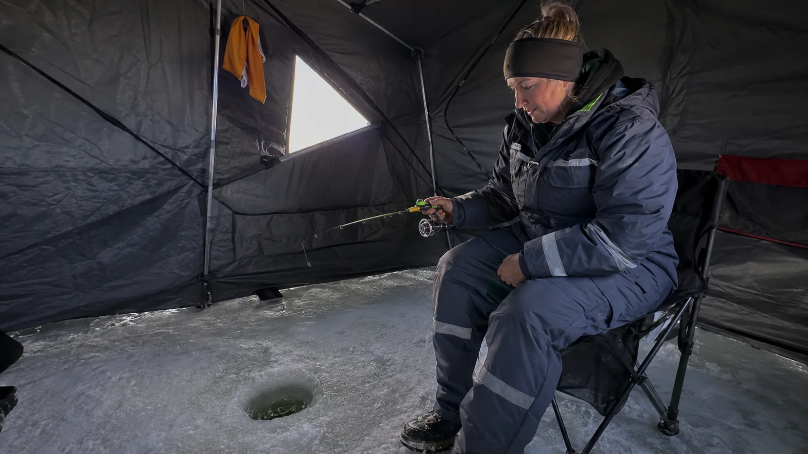 Bundled angler holding a short fishing rod inside an ice shelter.